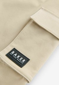 Beige stof met een gladde textuur, voorzien van een zwart rechthoekig etiket met de tekst "BAKER BY TED BAKER" in witte letters.