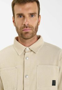 Beige corduroy jas met een gekraagde halslijn, zilveren knopen, twee borstzakken en een klein logo-applicatie op de linker zak.