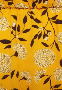 Gult tyg med en texturerad yta som har vita blommönster och bruna blad, vilket skapar en livlig och färgstark design.