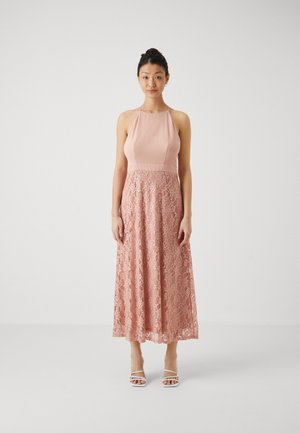 Robe maxi avec un corsage en satin rose et une jupe en dentelle. Elle présente un col halter, un motif de dentelle texturé et des couches transparentes. Sandales blanches à bout ouvert.
