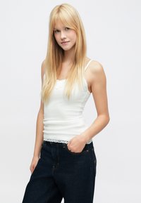 Hvit singlet med blonderkant nederst, kombinert med mørkeblå høytlivsjeans. Modellen står med hendene i lommene og håret løst.