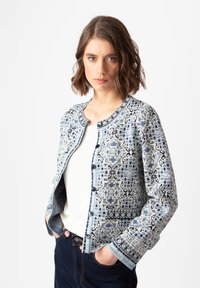 Mėlyna raštuota striukė su apvaliu kaklu, mygtukų uždarymu ir sudėtingais raštais. Yra tekstūrinis audinys ir kontrastingos apdailos detalės.