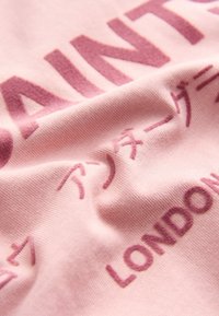 Vaaleanpunainen puuvillakangas, jossa kohokuvioituna tummanpunaista tekstiä eri kielillä. Sileä pinta, hieman ryppyinen ulkonäkö.