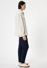 Lichtbeige shirt met een zak, gecombineerd met donkerblauwe broek en witte sneakers met bruine accenten. Glad materiaal en een ontspannen pasvorm.