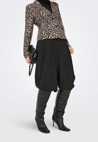 Femme portant un blazer imprimé léopard, un pantalon noir à jambes larges, des bottes en cuir noir montant jusqu'au genou, tenant un sac à main noir avec une anse tressée.