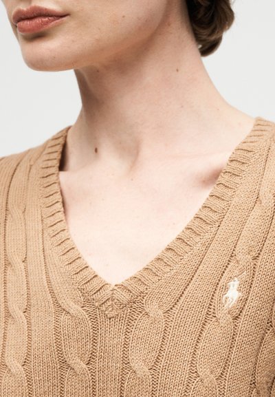 Κοντινή λήψη ενός ατόμου που φοράει μπεζ πουλόβερ V-λαιμόκοψη με πλεξούδα και ένα μικρό λευκό κεντημένο λογότυπο στο στήθος.