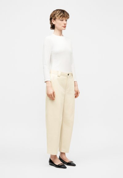 Femme debout portant un haut blanc à manches longues, un pantalon large taille haute beige et des ballerines noires à bouts pointus sur fond blanc.