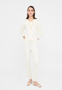 Il completo da loungewear color crema include una giacca con zip e colletto e pantaloni con coulisse, abbinati a scarpe piatte beige con texture.