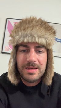 Faux pelslue med øreflippar, beigefarge, med myk tekstur og tykk, lodden design. Mørk genser i bakgrunnen.