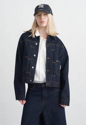 Femeie blondă purtând o jachetă și pantaloni din denim închis la culoare, cămașă albă și șapcă de baseball în dungi fine închisă, pe un fundal simplu, deschis la culoare.