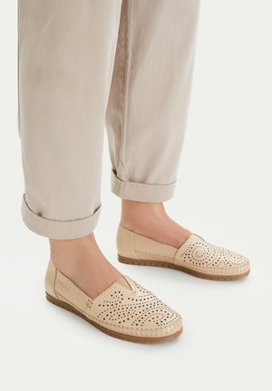 Mocasines beige perforados sin cordones, usados con pantalones doblados de color marrón claro, mostrados sobre un fondo blanco en un entorno casual.