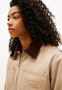 Chaqueta beige claro con cuello de pana marrón, cierre de cremallera en el frente y dos bolsillos en el pecho con logotipo bordado. Textura suave y diseño casual.