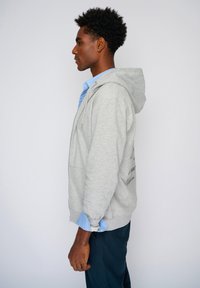 Felpa grigia con cappuccio, dotata di zipper frontale e tasche. Indossata sopra una camicia a colletto azzurro, esposta su uno sfondo semplice.