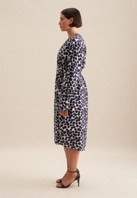 Abito a maniche lunghe con un motivo a pois blu navy e bianchi, vita cinte e silhouette fluida, abbinato a sandali neri con tacco alto.