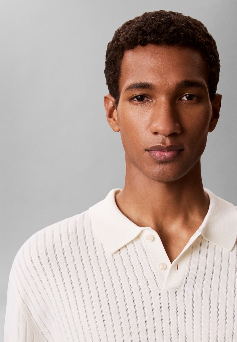 Jeune homme aux cheveux courts et bouclés portant une chemise blanche côtelée à col, face à un fond gris uni.