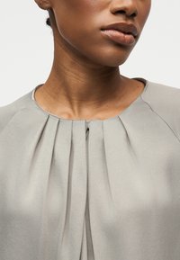 Gros plan d'une personne portant un chemisier gris clair avec un devant plissé et un col rond, montrant le cou et le bas du visage.