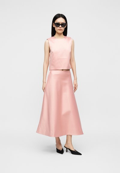 Γυναίκα που φοράει αμάνικο ανοιχτό ροζ τοπ και μακριά φούστα στο ίδιο χρώμα, μαύρα ψηλοτάκουνα mules και μαύρα γυαλιά ηλίου, στέκεται σε ανοιχτό φόντο.