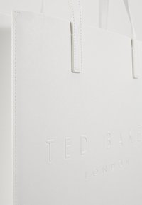 Sac fourre-tout blanc en matériau texturé, portant un logo en relief "Ted Baker London" et des poignées cousues. Design simple et rectangulaire.