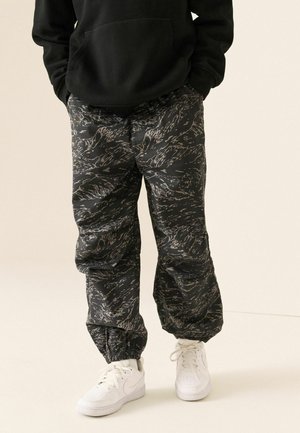 Person trägt schwarze und graue Camouflage-Hose mit elastischen Bündchen, weiße Sneaker und einen schwarzen Hoodie mit Händen in den Taschen.