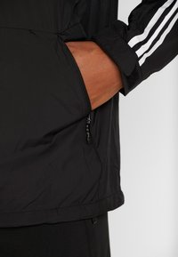 Svart vindjacka med dragkedjeficka och vita randdetaljer. Har en slät yta och en avslappnad passform med justerbara ärmslut.