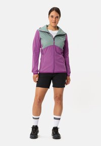 Lila und hellgrüne Zip-Jacke mit Kapuze, farblich abgesetzt, aus leichtem Material und mit zwei seitlichen Taschen, kombiniert mit schwarzen Shorts.
