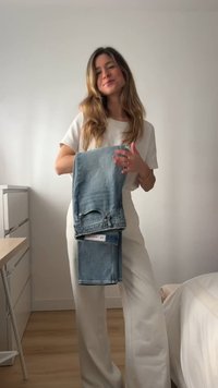 Valge riietusega naine hoiab volditud siniseid teksaseid ja seisab minimaalselt sisustatud toas, millel on puidust põrand ja valged seinad.