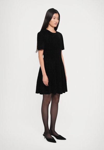 Young woman standing in a black short-sleeve dress, sheer black tights, and black pointed slingback heels on a plain white background.