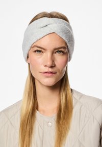 Una fascia per capelli grigia chiaro a maglia presenta un design attorcigliato, realizzata in materiale morbido. Il modello ha capelli lunghi e lisci e indossa una giacca trapuntata beige.