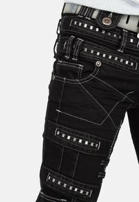 Pantaloni neri con molteplici tasche, cuciture bianche a contrasto e inserti metallici quadrati. Presentano un design patchwork unico e un tessuto resistente.