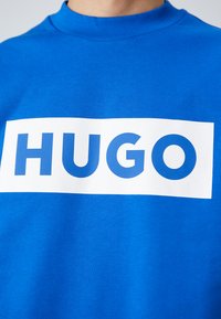 Blauwe sweater met een hoge ribbelhalslijn en gedurfde witte "HUGO" tekstlogo in een rechthoekig blokontwerp op de voorkant. Zachte stoftextuur.