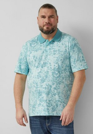 Mann mit Bart, der ein hellblaues Poloshirt mit floralem Muster und dunkelblaue Jeans trägt, steht vor einem schlichten hellen Hintergrund.