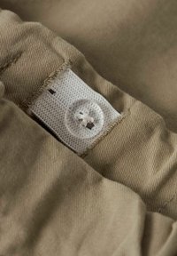 Närbild av khaki-tyg med en vit etikett och en knapp. Materialet är texturerat och något rynkat, med synliga sömnadsdetaljer.