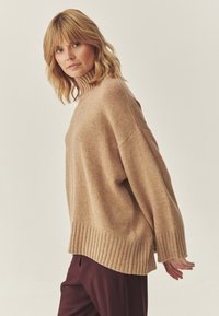 Maglione lavorato a maglia beige con collo alto, orlo a coste e design oversize. Indossato con pantaloni bordeaux scuri. La texture morbida è visibile.