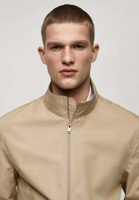 Beige lynlåsjakke med opretstående krave. Glat tekstur og minimalistisk design, der har en sølvlynlås foran og ingen synlige mønstre.
