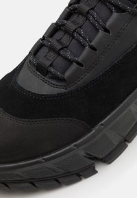 Chaussure de sport noire composée d'un mélange de suède et de mesh, avec un embout renforcé, une semelle épaisse et texturée, ainsi qu'un système de laçage contrasté.