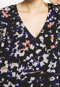 Haut en tissu noir avec un décolleté en V, présentant un motif floral en violet, blanc et corail. Texture légère et lisse.