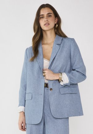 Femme aux longs cheveux bruns portant un blazer bleu clair et un pantalon assorti, une blouse blanche, des boucles d'oreilles créoles dorées, des bagues et des bracelets.