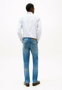 Jeans di denim blu con una finitura scolorita, indossati con una camicia a righe blu chiaro e bianco, visti da dietro, che mostrano la vestibilità e i dettagli delle tasche.