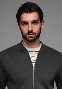 Chaqueta gris con cremallera y cuello redondo, con una cremallera plateada y bordes acanalados, llevada sobre una camiseta a rayas en color crema y negro.