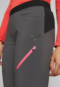 Šedé sportovní kalhoty s černým pasem, vybavené růžovou kapsou na zip a logem u stehna. Hladká látka s přiléhavým střihem.