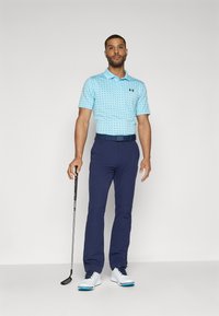 Polo de culoare albastru deschis cu un model cu puncte, asortat cu pantaloni negri și pantofi de golf albi, ținând un club de golf.
