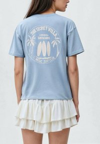 Modrá bavlněná tričko s bílou grafikou surfovacích prken a palem. Spárováno s vrstvenou bílou sukní s volány.