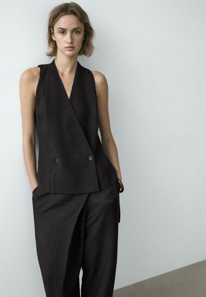 Giovane donna con capelli corti che indossa un gilet nero doppiopetto senza maniche e pantaloni larghi abbinati, mani nelle tasche, sfondo neutro.