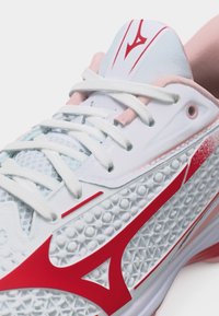 Κοντινή λήψη λευκού και ροζ αθλητικού παπουτσιού με υφασμένη σόλα και εμφανές κόκκινο λογότυπο στο πλάι και στη γλώσσα, δεμένο με λευκά κορδόνια.