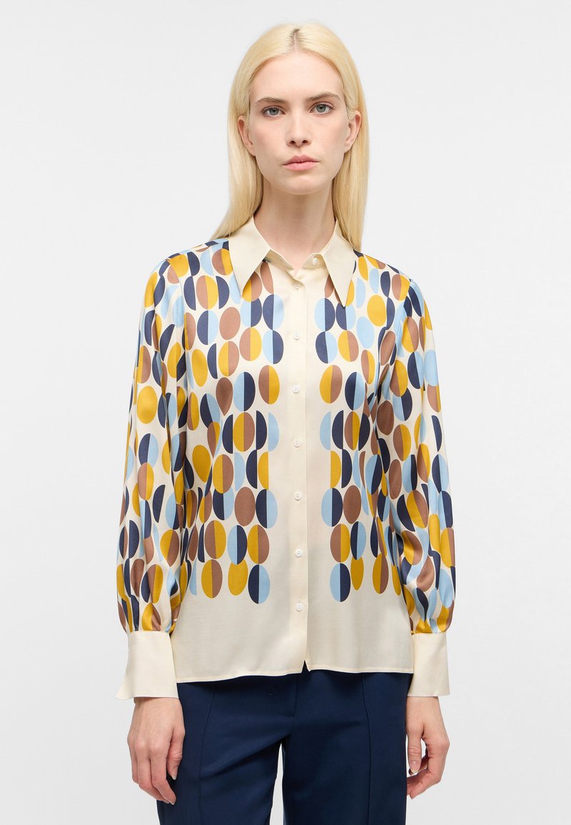 Blusa a maniche lunghe in tessuto crema, con un motivo geometrico di ellissi blu, marroni e gialli. Colletto a V e chiusura con bottoni.
