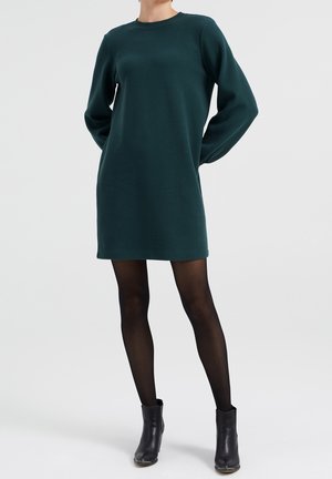 Vrouw die een donkergroene, lange mouwen, getextureerde jurk draagt, zwarte doorschijnende panty's en zwarte enkellaarzen, staand met haar handen achter haar rug.