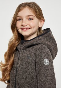 Giacca con cappuccio e zip in tessuto strutturato grigio scuro, caratterizzata da un'applicazione decorativa sul manica sinistra e un rivestimento nero liscio all'interno del cappuccio.