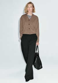 Cardigan di lana marrone con bottoni neri, indossato sopra un top grigio. Pantaloni neri a vita alta e una borsa a tracolla in pelle nera.