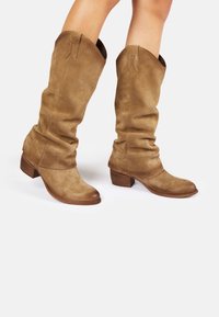 Bottes en daim marron à mi-mollet avec un design loose, bout arrondi et un petit talon empilé. Détails de couture au niveau du bout et texture douce.