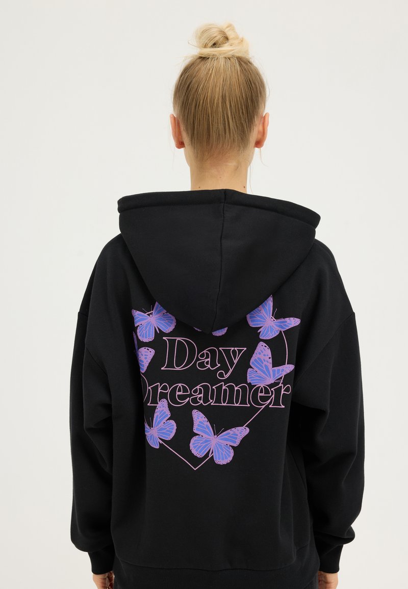 Personne blonde avec les cheveux en chignon, portant un sweat à capuche noir avec des papillons violets et le texte "Day Dreamer" au dos, sur un fond uni.
